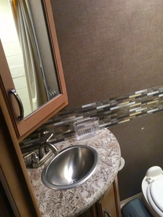 RV rental bathroom features: compact stainless steel sink, speckled countertop, tiled accent wall, and part of a white toilet.. Thor Freedom Elite 2018
