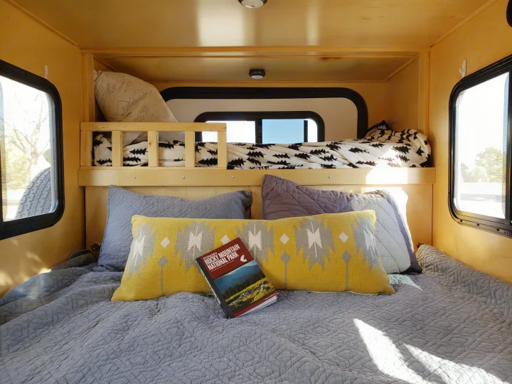 Cozy RV rental interior shows a lower bed with a national park guide, patterned pillows, and a raised bunk bed. Perfect for trips!. Escapade Ridge 2018