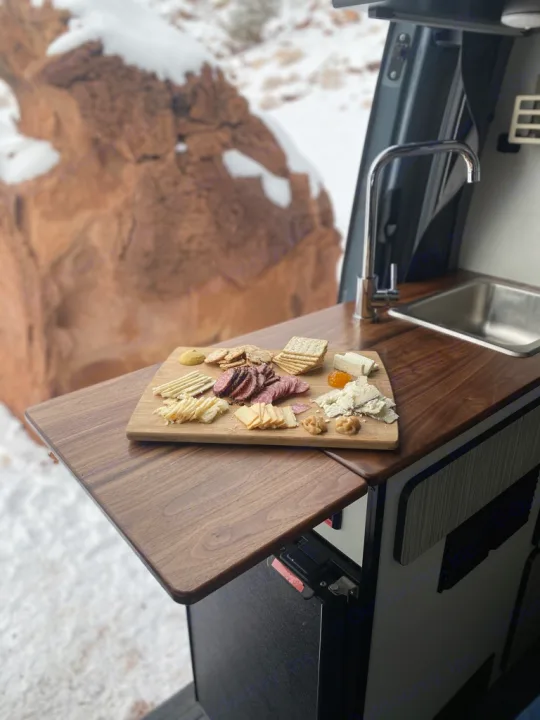 Inside an rv rental, a charcuterie board sits on a wooden pull-out table. Snowy landscape visible outside, perfect winter dining.. Winnebago Revel 2021