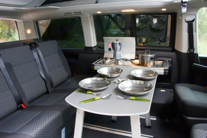 Inside an RV rental: a compact camper van kitchen with a stove, sink, coffee maker, and dining table set with plates.. Volkswagen Transporter California Beach 2019