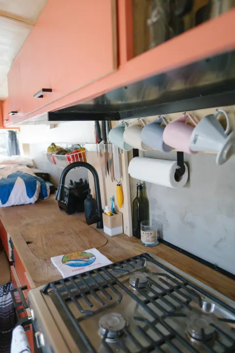 An RV rental's cozy kitchen area with vibrant orange cabinets, wood countertop, sink, stove, and organized hanging mugs.. Ford Transit 2015