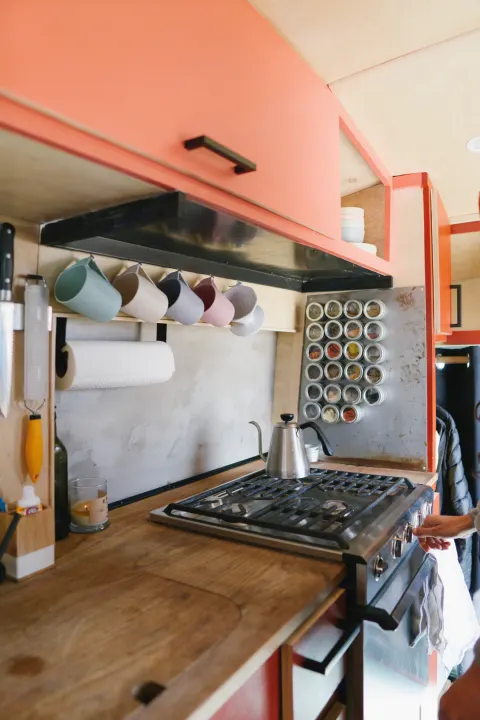 Compact RV rental kitchen featuring a gas stove, wood counters, magnetic spice rack, and colorful hanging mugs.. Ford Transit 2015