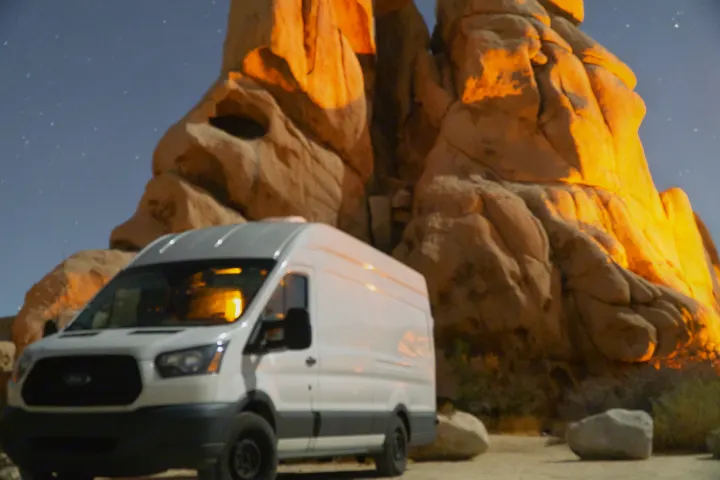 White RV rental van parked at night against majestic, orange-lit rock formations under a sky with visible stars.. Ford Transit 2015