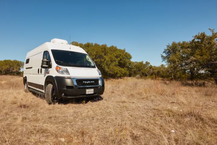 A white RV rental camper van sits in a golden, sun-drenched field with green trees in the distance under a clear blue sky.. Dodge 2500 Promaster 2019