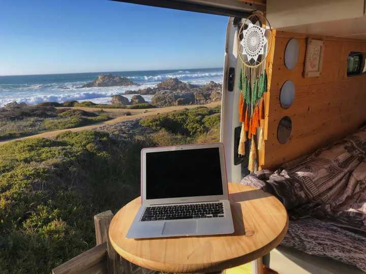 RV rental: A laptop on a wooden table overlooks a scenic rocky coast with crashing waves from inside a camper van.. Ford Transit 2015