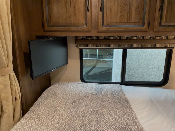 Cozy RV rental bedroom features a patterned blanket, wall-mounted TV, wooden cabinets, and window. Ideal for travel comfort.. Coachmen Prism 2019