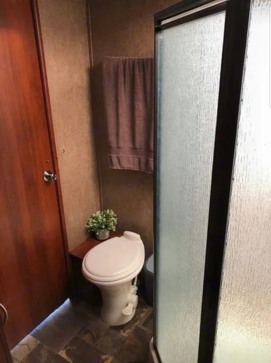 Compact RV rental bathroom featuring a toilet, decorative plant, brown towel, and textured glass shower enclosure.. Forest River Crusader 2015