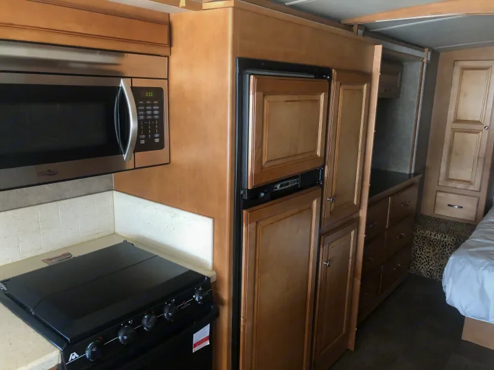 Inside an RV rental, showing a full kitchen with microwave, stove, refrigerator, wood cabinets, and nearby sleeping area.. Itasca Sunstar 2015