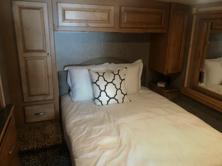 Inside a cozy RV rental: a comfortable bed with white linens, surrounded by wood cabinets & storage.. Itasca Sunstar 2015