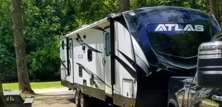 A large, white and black Atlas fifth-wheel RV is parked at a sunny, treed campsite, ideal for an rv rental.. Dutchmen Other 2020