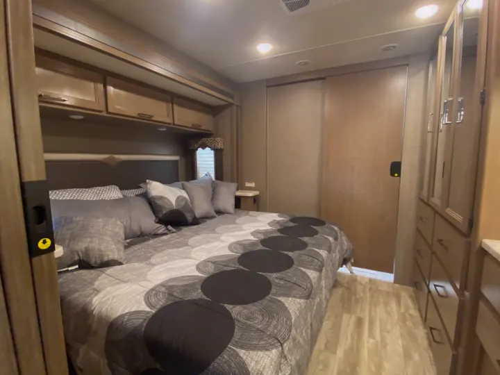 Cozy RV rental bedroom with patterned comforter, overhead storage, and mirrored wardrobe for your travels.. Thor Motor Coach Hurricane 2021