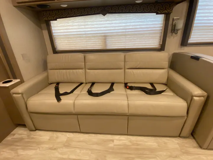 This RV rental boasts a comfortable three-person beige sofa with seatbelts, window blinds, and overhead storage for travel.. Thor Motor Coach Hurricane 2021