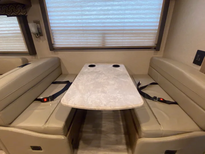 RV rental dinette setup: a light-colored table with cup holders, two bench seats with seatbelts, and pleated window blinds.. Thor Motor Coach Hurricane 2021