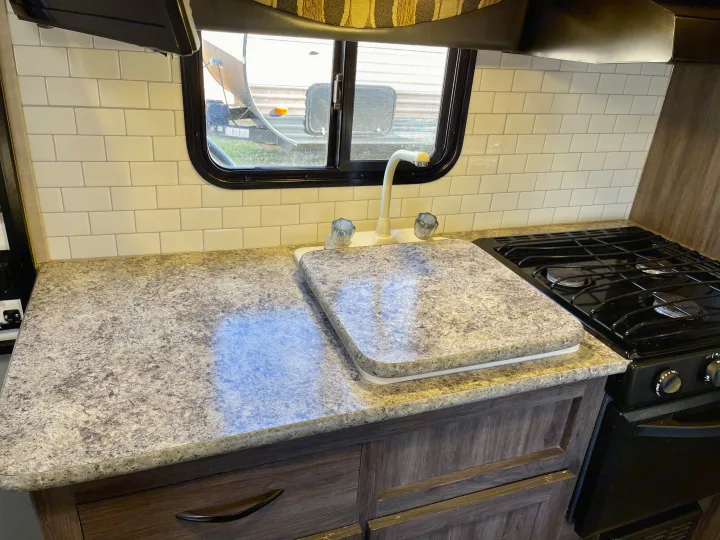 A modern RV rental kitchen featuring a speckled counter, white subway tile backsplash, sink with faucet, and gas stovetop.. Pacific Coachworks Panther 2019