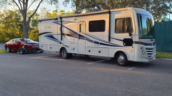 White Fleetwood Flair motorhome (RV rental) towing a red car on a dolly, parked in an asphalt lot.. Fleetwood Flair 2017