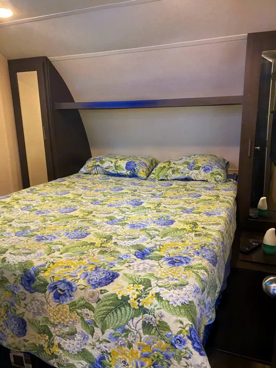 Cozy RV bedroom for your next RV rental adventure. Features a comfortable bed with floral bedding and built-in storage.. Forest River Wildwood 2018