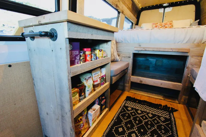 RV rental interior: Built-in wood shelves stocked with food, a cozy bed, and a patterned rug for comfortable mobile living.. Mercedes-Benz Sprinter 419 LWB High Roof 2020