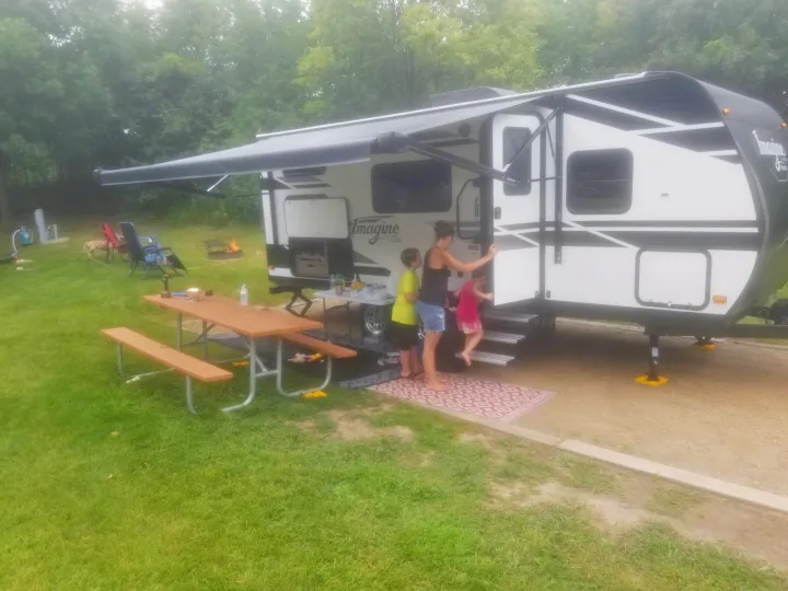Family at a spacious RV rental campsite. Features an Imagine XLS travel trailer with awning, picnic table, fire pit, and dog.. Grand Design Imagine XLS 2021