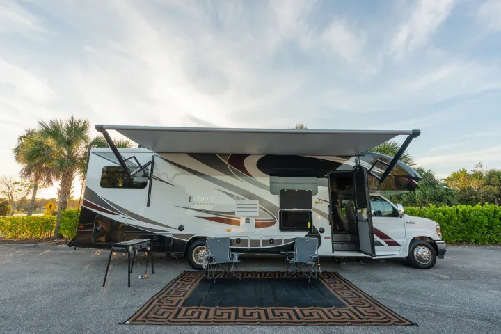 This RV rental is set up for outdoor living with an extended awning, chairs, mat, and grill on an asphalt pad.. Entegra Coach Esteem 31F 2021