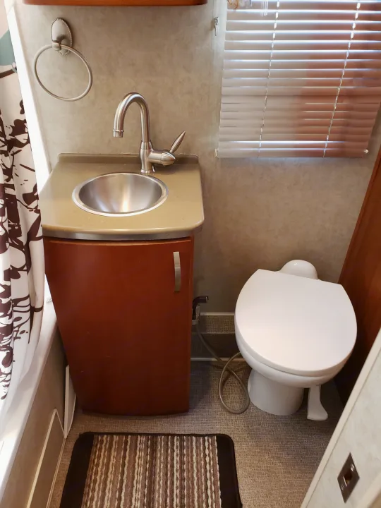 RV rental bathroom with a compact sink, vanity, toilet, window blinds, and shower curtain. A bidet sprayer is near the toilet.. Fleetwood Pulse 2009