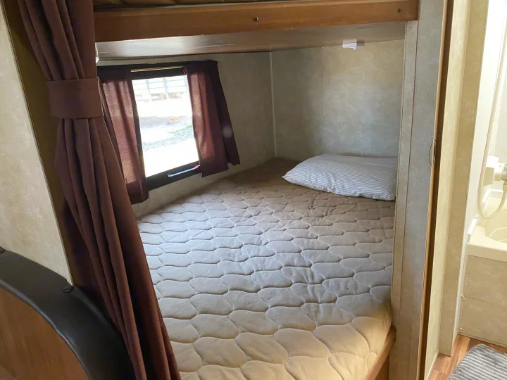 Inside an RV rental, a compact sleeping area features a quilted bed, striped pillow, and window with brown curtains.. Gulf Stream Kingsport 2015