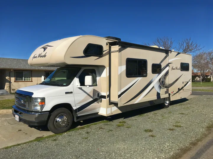 A beige and white Four Winds Class C RV rental, featuring black and white striped decals, is parked on a sunny gravel driveway.. Thor Motor Coach Four Winds 2018