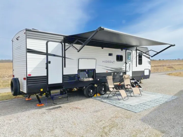 This RV rental travel trailer is set up on gravel, ready for outdoor enjoyment with an awning, outdoor kitchen, and chairs.. Keystone Hideout 2021