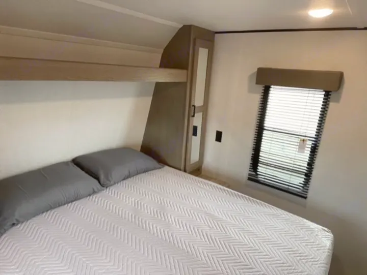 Spacious RV rental bedroom featuring a comfy bed with grey pillows, overhead bunk, wardrobe, and window with blinds.. Keystone Hideout 2021