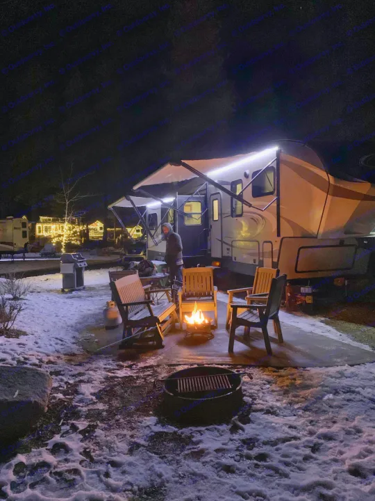 Cozy winter night at an **RV rental** campsite. A person, illuminated RV, campfire, and chairs are on snow.. Keystone Cougar 336BHS 2018