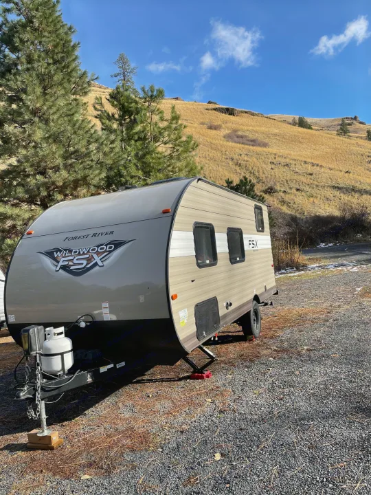 A Forest River Wildwood FSX RV rental is set up for camping on a gravel lot, backed by pine trees and golden hills under a clear sky, offering an ideal outdoor escape.. Forest River Wildwood 2019