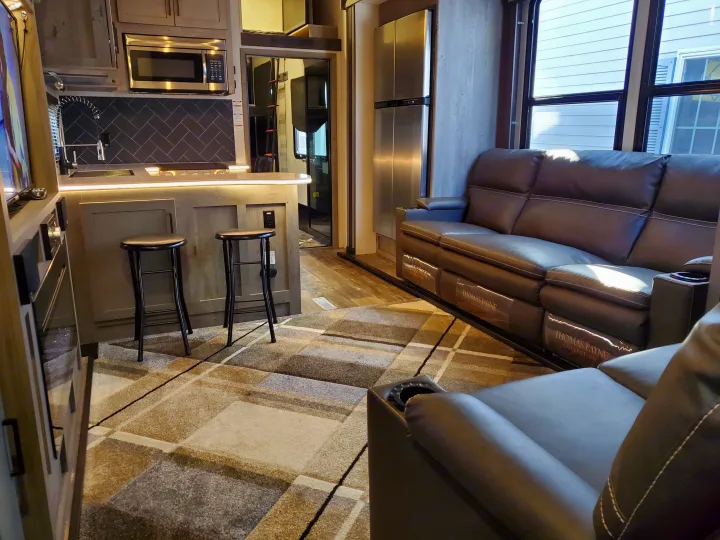 Spacious rv rental interior features a modern kitchen with microwave/sink, bar stools, and a plush leather sofa with cup holders.. Keystone Raptor 2020