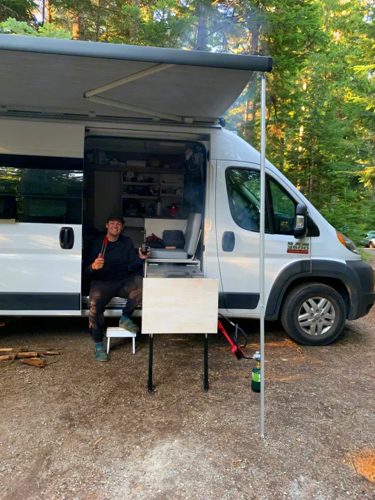 Enjoying an RV rental adventure! A man smiles, cooking outside his white campervan under an awning in a forest.. RAM ProMaster 3500 2017