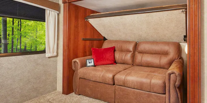 RV rental interior: a cozy brown sofa with red pillow, a hidden fold-down bed above, and a beautiful forest window view.. Jayco Eagle HT 2018