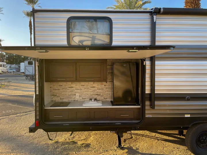 Equipped outdoor kitchen of an RV rental with sink, stove, and fridge, perfect for camping. Palm trees visible in sunny background.. Forest River Cruise Lite 2020