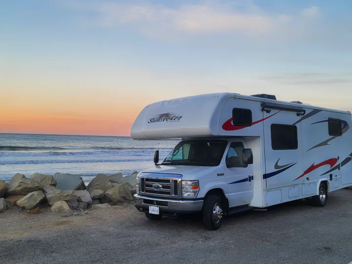An RV rental, a Sunseeker motorhome, is parked by the ocean at sunset, ideal for a scenic beach camping trip.. Forest River Sunseeker 2019