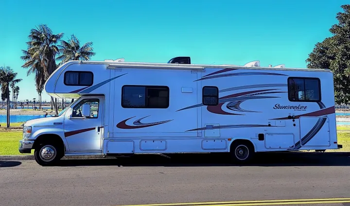 A white Sunseeker Class C RV parked by the water with palm trees under a clear sky, perfect for your next rv rental adventure.. Forest River Sunseeker 2015