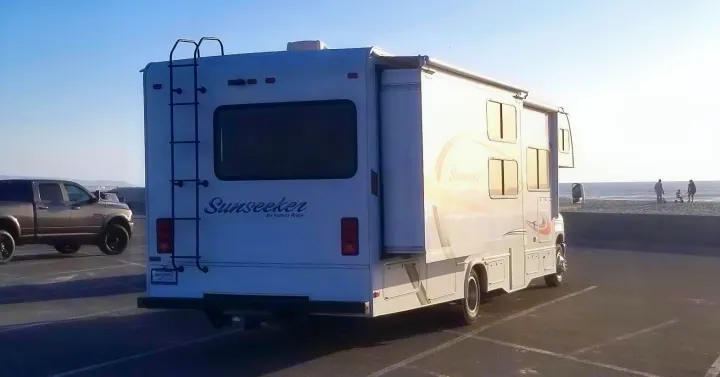 Rear view of a white Sunseeker RV rental parked at a sunny beach lot, with a pickup truck and ocean in the background.. Forest River Sunseeker 2015