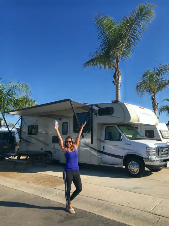 Happy woman in blue tank top raises arms by her RV rental at a sunny palm-tree lined campsite, holding a water bottle.. Ford Coachman 2019