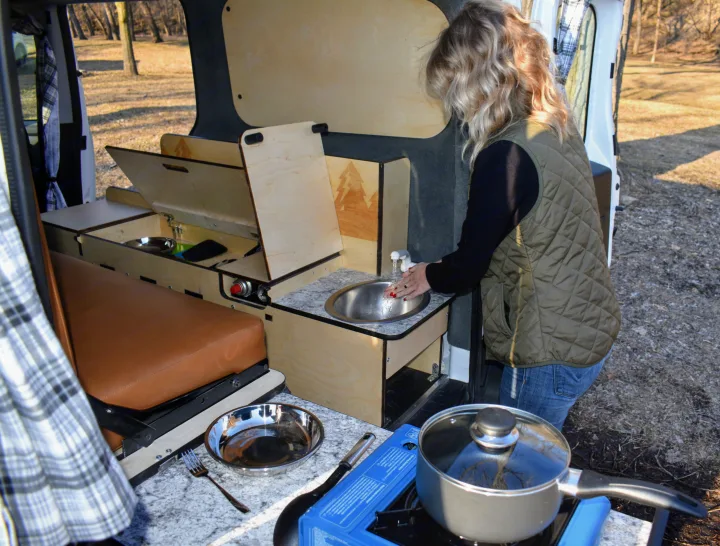 Woman washes hands in her camper van's compact kitchen. This rv rental offers a sink, stove, and seating for outdoor living.. Dodge Promaster City 2020