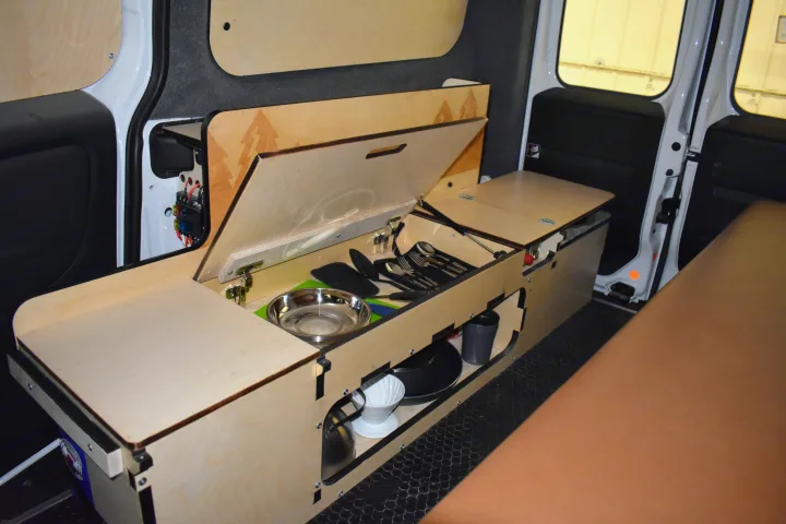 Compact RV rental interior featuring a modular wooden kitchen unit with opened lid showing cooking essentials, and a brown cushioned bed.. Dodge Promaster City 2019
