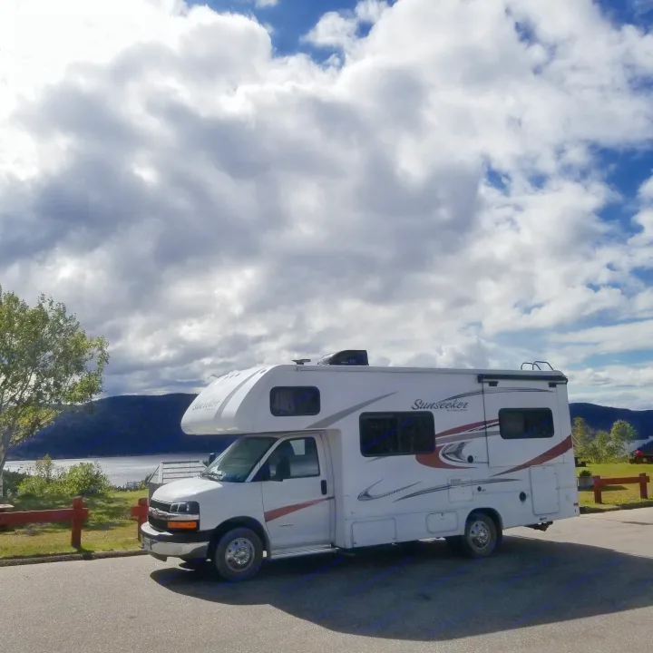 White Sunseeker RV rental parked by a lake, mountains, and a partly cloudy sky.. Forest River Sunseeker 2016