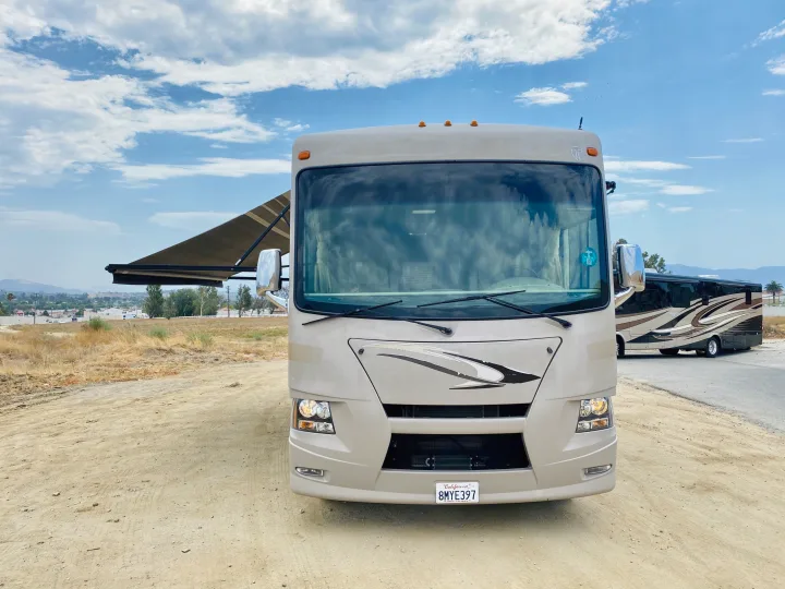 Front view of a beige RV rental parked on a dirt lot with its awning out, another RV behind, under a cloudy sky.. Thor Motor Coach Windsport 2014