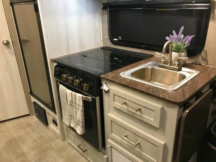 Compact RV rental kitchen with a stainless steel sink, black stove, refrigerator, and light wood cabinets.. Forest River Rockwood 2021