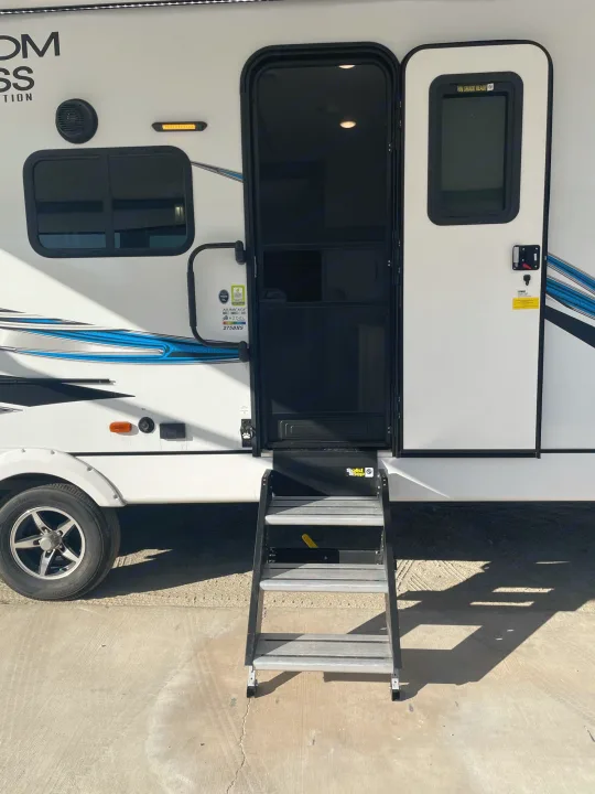 White travel trailer RV rental, with its entry door open, screen visible, and extended aluminum steps leading to the ground.. Coachmen Freedom Express 2021