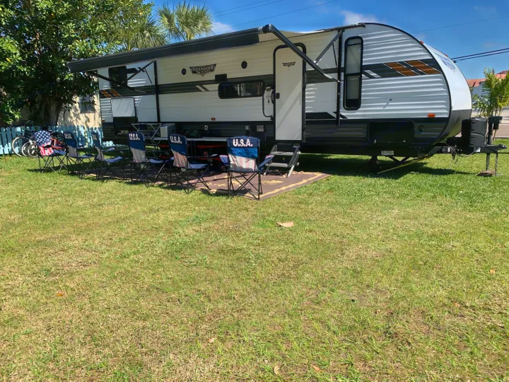 Enjoy an RV rental: a white travel trailer with an extended awning, USA chairs, and a table on a sunny grassy site.. Forest River Wildwood X-Lite 2020