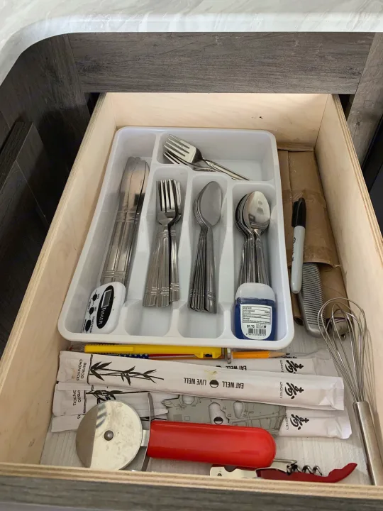 In an rv rental, an open drawer shows a utensil organizer with silverware, a timer, hand sanitizer, pizza cutter, and whisk.. Forest River Wildwood X-Lite 2020