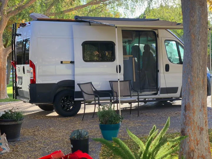 RV rental: White camper van with an extended awning, chairs, and potted plants creates a cozy outdoor living space.. Dodge RAM Promaster 2500 159' Wheelbase 2017