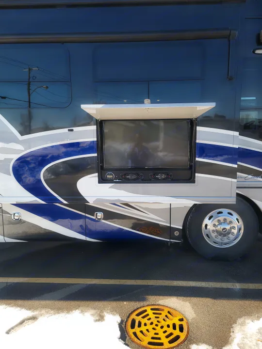 A Holiday Rambler RV rental, painted in blue, grey, and white, with an open compartment revealing an outdoor TV and speakers.. American Revolution 2019
