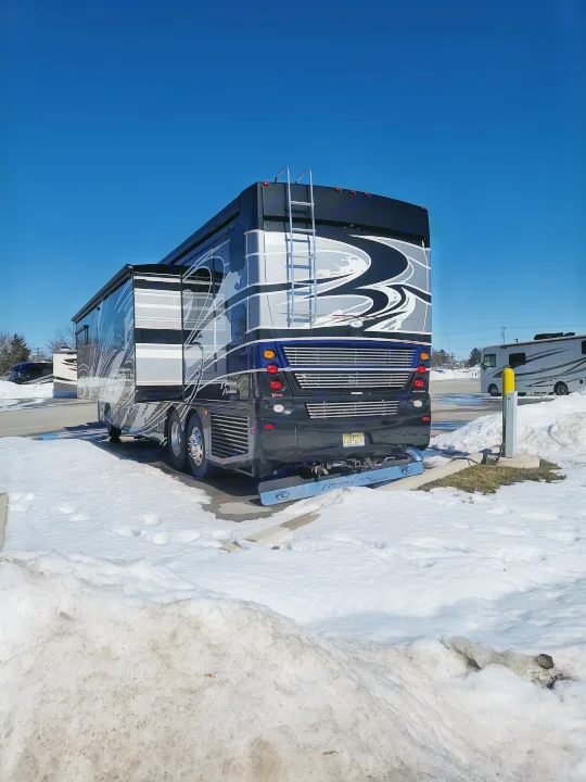 Large black American Coach Revolution RV parked in snow at a winter campground. Perfect for an RV rental adventure under a clear blue sky!. American Revolution 2019