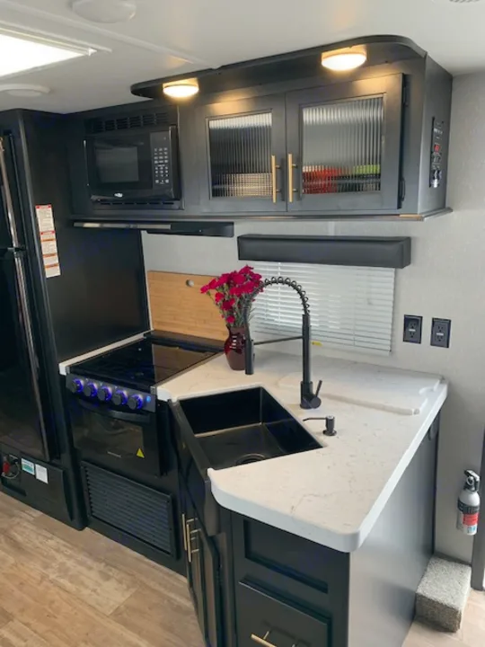 Modern RV rental kitchen interior featuring sleek dark cabinets, a white counter, sink, microwave, and stovetop/grill.. Cherokee Grey Wolf 27DBH 2021